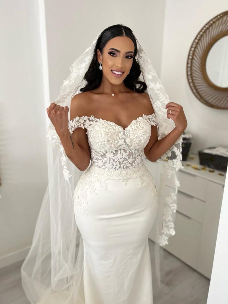 Mariee en robe sirene a dentelle florale et epaules denudees, ecartant son long voile brode, le matin des preparatifs