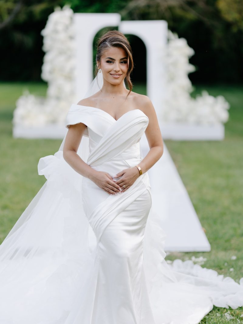 Mariee seule sur une pelouse, robe drapee a longue traine, devant les arches blanches de la ceremonie