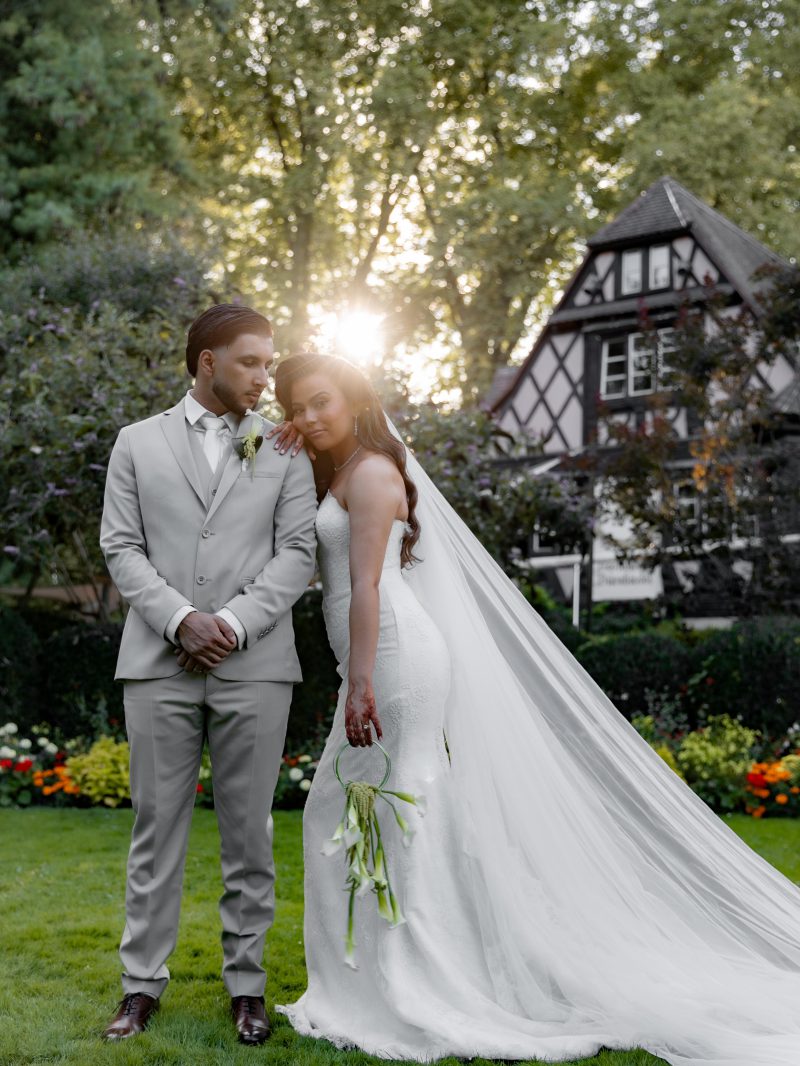 Mariee et marie enlaces dans un jardin au soleil couchant, robe sirene en satin ivoire a longue traine, devant une maison a colombages