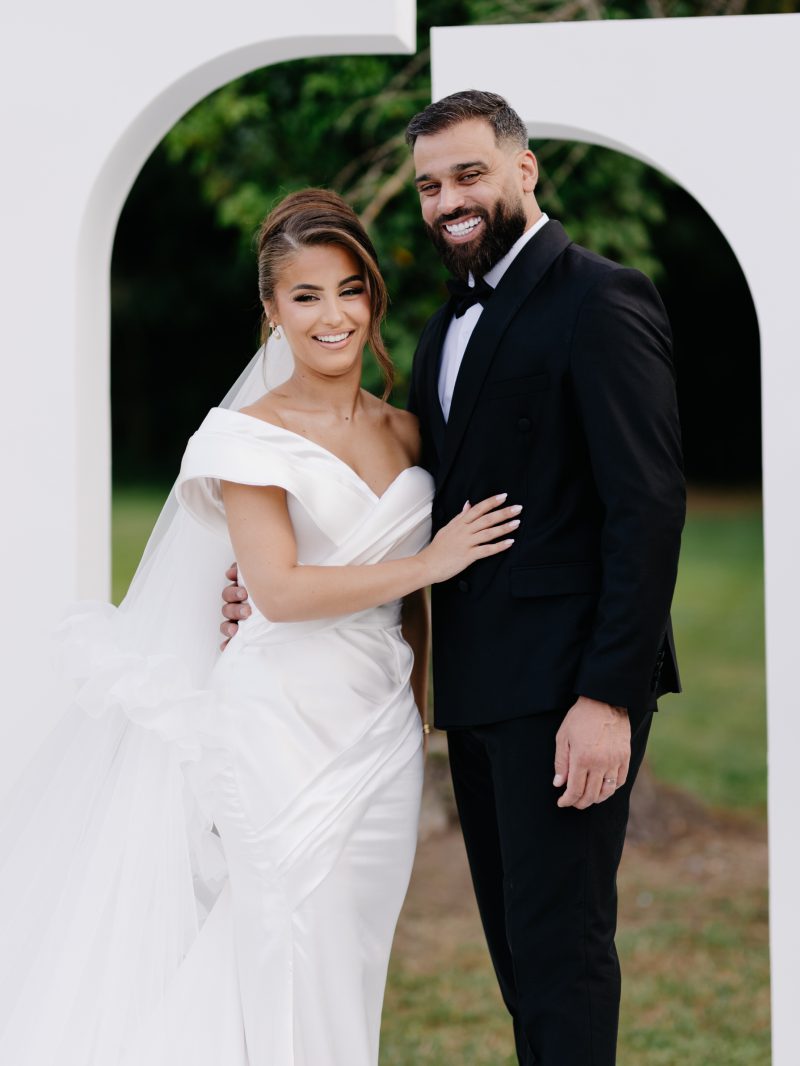 Mariee en robe drapee a epaule denudee aux cotes de son mari en smoking noir, devant une arche blanche en exterieur